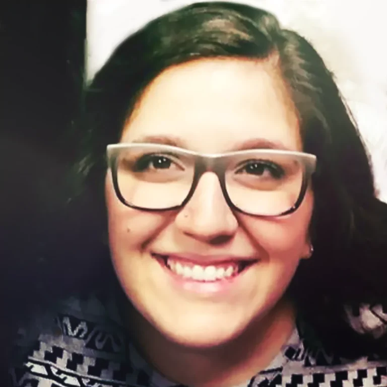 Smiling woman with glasses and dark hair, wearing a patterned top.